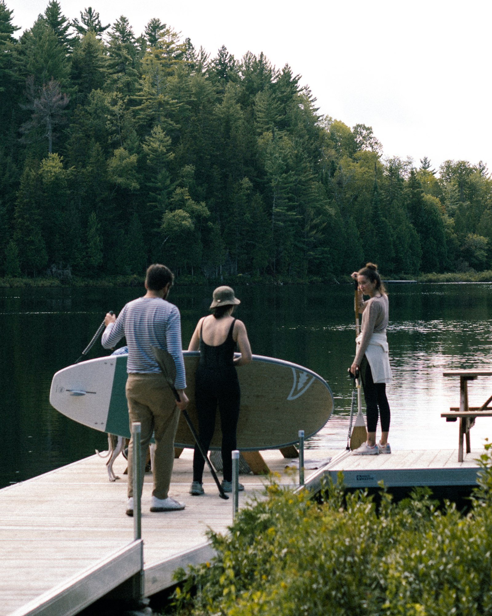 Three friends on a lake dock with a paddleboard and paddles, pine forest across the water