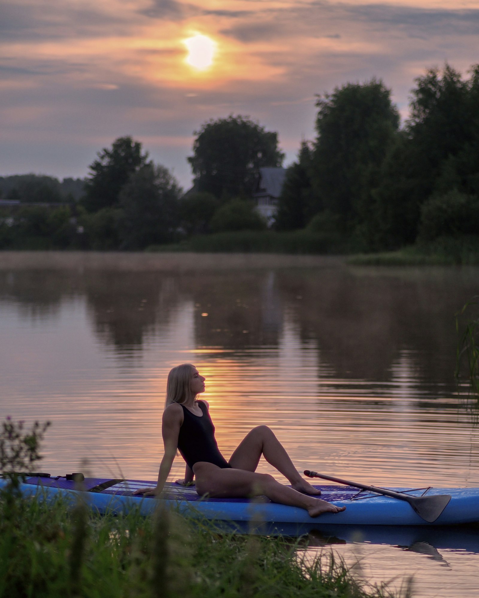 A woman in a swimsuit sits back on a blue paddleboard at the lake's edge, eyes closed, facing the low sun